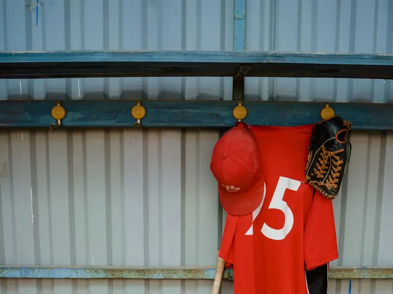 Red baseball jersey, cap and glove hanging in a dugout above a bat and balls